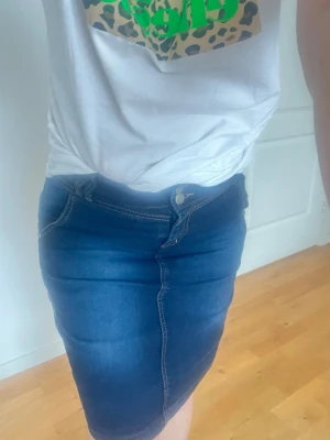 Mörkblå jeanskjol med fickor - Snygg mörkblå jeanskjol i klassisk modell med framfickor och synliga sömmar. Kjolen har knapp- och dragkedjestängning framtill och är lätt figurnära. Perfekt att matcha med en t-shirt eller hoodie för en avslappnad look.