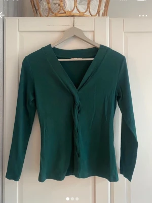 Mörkgrön långärmad tröja - Mörkgrön långärmad tröja med v-ringad hals och en diskret twistad detalj framtill. Snygg och enkel modell som passar till mycket. Tillverkad i mjukt material och har en normal passform. Strl. M.