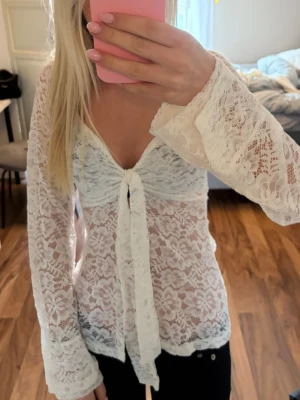 Vit spetsblus med knyt framtill design by si - Säljer en superfin vit blus i spets med långa ärmar och v-ringning. Blusen har ett snyggt knyt framtill och är helt genombruten med blommigt spetsmönster. Perfekt för dig som vill ha en romantisk och trendig look.