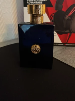 Versace blå parfymflaska - Elegant parfymflaska från Versace i mörkblått glas med fyrkantig form och guldfärgat lock. Flaskan har en guldig Versace-medusa på framsidan som ger en lyxig känsla. Perfekt för dig som gillar exklusiva dofter och snygg design.