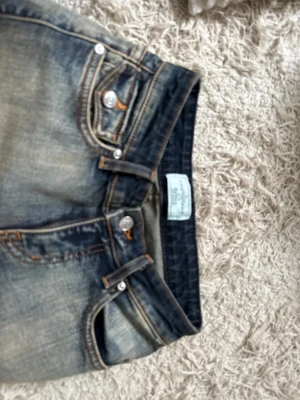 Blå bootcut jeans från Replay - Säljer ett par blå bootcut jeans från Revice med slitningar och kontrastsömmar. Jeansen har två fickor fram och två bak med lock och knapp, samt detaljerade nitar. Klassisk femficksmodell i denim med snygg tvätt.  