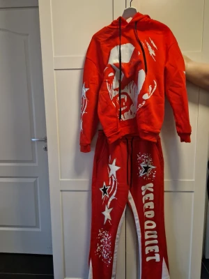 Rött loungewear-set med tryck - Säljer ett rött loungewear-set med hoodie och byxor. Hoodien har ett stort vitt tryck av ett ansikte och svarta dragsnören. Byxorna har vita stjärnor, texten 'KEEP QUIET' och grafiska detaljer. Setet är gjort i mjukt material, perfekt för chill dagar hemma. Kan köpas separat också