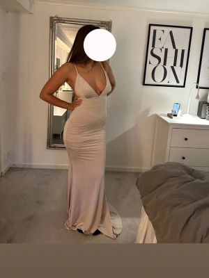 Ljus långklänning med öppen rygg - Elegant långklänning i ljus beige ton med djup v-ringning och tunna axelband. Klänningen har en figurnära passform och en snygg öppen rygg. Perfekt för speciella tillfällen där du vill sticka ut.