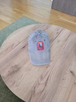 Grå keps från Parajumpers - Snygg grå keps från Parajumpers med broderad patch framtill i rött och blått. Kepsen är tillverkad i 100% bomull och har klassisk böjd skärm samt justerbar rem bak. Perfekt accessoar för en avslappnad och sportig stil.