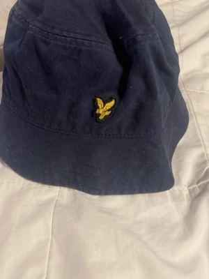 Mörkblå bucket hat från Lyle & Scott - Snygg mörkblå bucket hat från Lyle & Scott med klassisk gul broderad logga framtill. Hatten är i bomull och har en avslappnad passform. Perfekt accessoar för att lyfta din stil och skydda mot solen.