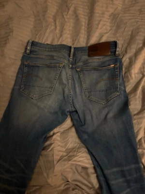 Blå jeans från G-Star RAW - Säljer ett par blå jeans från G-Star RAW med klassisk femficksdesign och snygga slitningar på framsidan. Jeansen har en rak passform och detaljerade sömmar på bakfickorna. Perfekta för en avslappnad och trendig look.