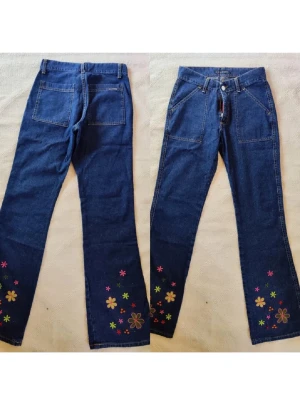 Blå jeans med broderade blommor - Unika blå jeans med utsvängda ben och färgglada broderade blommor längst ner på båda benen. Jeansen har två stora bakfickor och framfickor, samt klassisk knappgylf. Perfekta för dig som vill sticka ut med en retro och lekfull stil.