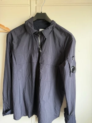 Mörkblå overshirt från C.P. Company - Stilren mörkblå overshirt från C.P. Company med klassisk krage, dragkedja framtill och ikonisk linsdetalj på ärmen. Tillverkad i lätt, tekniskt material som ger en modern look. Perfekt lager-på-lager-plagg för dig som gillar streetwear och funktion.