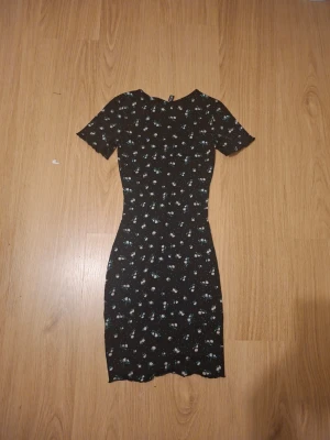 Svart blommig kortklänning - Söt svart kortklänning med små blommor i vitt och blått. Klänningen har korta ärmar och en figurnära passform som framhäver siluetten. Perfekt för sommarens alla tillfällen och enkel att styla med sneakers eller sandaler.