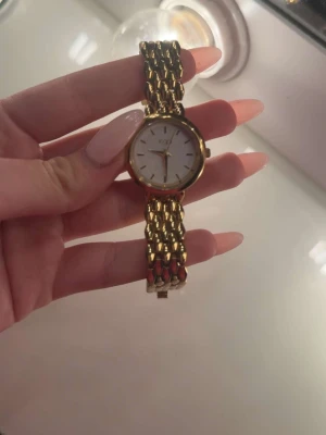 Guldig klocka från Regal - Stilren klocka från Regal med rund vit urtavla och guldfärgat armband i metall. Klassisk design med tydliga markeringar och elegant länkarmband som ger en lyxig känsla. Perfekt accessoar för att lyfta din outfit.