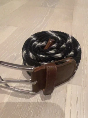 Svart och vitt flätat bälte - Snyggt flätat bälte i svart och vitt med bruna skinn-detaljer och silverfärgat spänne. Perfekt för att ge jeansen en cool touch. Bältet har en flexibel, vävd design som gör det enkelt att justera längden.