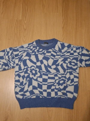 Blåvit stickad tröja med blommönster - Cool stickad tröja i blått och vitt med grafiskt blommönster och breda ribbade muddar. Tröjan har rund hals och är gjord i ett mjukt material som känns skönt mot huden. Perfekt för dig som gillar retrovibbar och vill sticka ut lite extra.