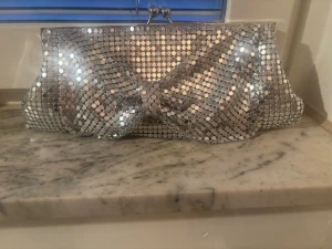 Silvrig glittrig kuvertväska - Säljer en silvrig kuvertväska med glittrande paljetter över hela väskan. Väskan har en klassisk avlång form och metallspänne upptill. Perfekt för dig som vill sticka ut med en riktigt snygg accessoar.
