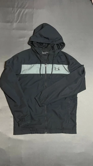 Svart vindjacka från Under Armour - Säljer en riktigt fräsch Under Armour jacka i mycket fint skick. Jackan är perfekt för både träning och vardagsbruk. Den har en klassisk svart design med en snygg grå panel över bröstet och den ikoniska loggan tydligt synlig. 
