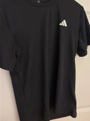 Svart Adidas t-shirt med logga - Säljer en svart t-shirt från Adidas med den klassiska vita loggan på bröstet. T-shirten har rund halsringning och korta ärmar. Perfekt för en sportig och avslappnad stil.