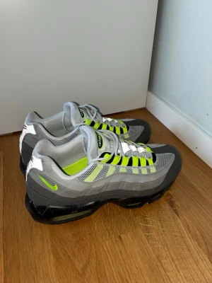 Nike Air Max 95 Neon  - Nike Air Max 95 sneakers med ikonisk neonfärg på snörning och detaljer. Skorna har en grå och svart färgskala med mesh och mocka-material, samt synlig Air Max-dämpning i sulan. Perfekt för dig som vill ha en klassisk och sportig look.
