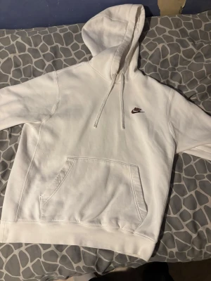 Vit hoodie från Nike med logga - Vit hoodie från Nike med broderad brun logga på bröstet. Klassisk design med stor huva, dragsnören och en rymlig magficka. Mjuk bomullskänsla och avslappnad passform, perfekt för chill dagar.