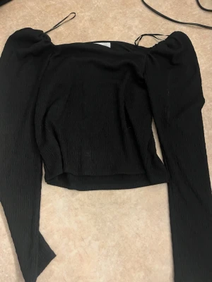 Svart långärmad croppad topp - Trendig svart croppad topp med långa ärmar och lätt puff vid axlarna. Toppen har en åtsittande passform och tunna axelband som ger en modern look. Perfekt att styla med höga jeans eller kjol.