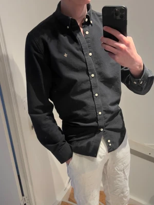 Svart skjorta från Morris - Klassisk svart skjorta från Morris med button-down krage och vita knappar. Skjortan har en liten broderad logga på bröstet och är till sopass enkel att soppa till jeans eller chinos. Tillverkad i mjuk bomull och har dsikret normal passform.