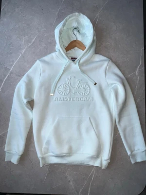 Vit hoodie med cykelmotiv Amsterdam - Snygg vit hoodie från Tommy Hilfiger med broderat cykelmotiv och texten 'AMSTERDAM' på bröstet. Hoodien har huva med dragsko och guldfärgade detaljer, samt en stor magficka. Perfekt för dig som gillar stilrena plagg med coola detaljer.