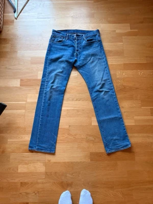 Levi’s 501 32/32 - Klassiska blå Levis jeans med rak passform och fem fickor modell 501. Jeansen har en knappgylf och bälteshällor. Materialet är denim i bomull och färgen är mellanblå med lätt slitning på låren. Perfekta för en avslappnad och tidlös stil. Storlek 32/32