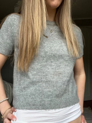 Grå kortärmad stickad tröja - Mysig grå stickad tröja med korta ärmar och rund hals. Tröjan har en enkel och stilren design med ribbade muddar vid ärmslut och nederkant. Perfekt att matcha med jeans eller kjol för en avslappnad look.