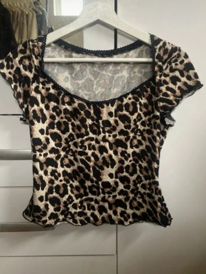 Leopardmönstrad topp med spetskant - Snygg leopardmönstrad topp med korta ärmar och dekorativ svart spetskant vid halsringningen. Toppen har en figurnära passform och är tillverkad i ett mjukt, stretchigt material som ger en bekväm känsla. Perfekt för dig som vill sticka ut med ett djurmönster.
