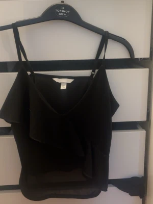 Svart volangtop från H&M - Säljer en svart linnetopp från H&M med tunna axelband och snygg volangdetalj framtill. Toppen har v-ringning och är perfekt för dig som gillar en enkel men ändå trendig look.
