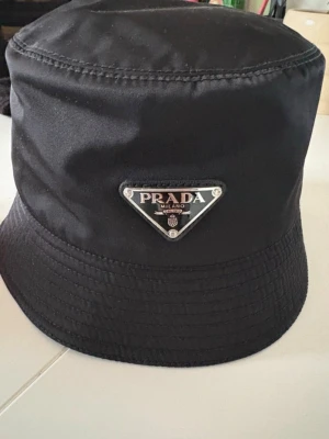 Prada Hatt - Svart bucket Hat från prada, klassiska triangulära logomärket för dig med klass.  *Äkta vara, kvitto medföljer* Pris kan diskuteras, tveka ej att skriva vid frågor!