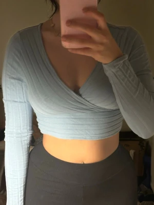 Ljusblå omlottopp med lång ärm - Trendig ljusblå croppad omlottopp med ribbad struktur och djup v-ringning. Toppen har långa ärmar och sitter tight över kroppen, perfekt för en snygg och modern look.