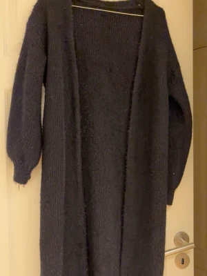 Lång svart stickad kofta - Mysig lång svart stickad kofta med öppen front och långa ärmar. Perfekt att slänga över en t-shirt eller hoodie för extra värme. Koftan har en avslappnad passform och är gjord i ett mjukt, fluffigt material.