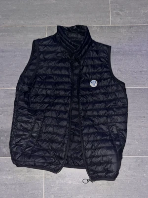 Svart dunväst från Save The Duck - Snygg svart dunväst från Save The Duck med quiltad design och hög krage. Västen har en rund logotyp på bröstet och dragkedja framtill. Perfekt för lager-på-lager och ger en sportig look.