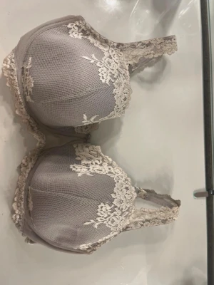 Grå spets-bh med vadderade kupor - Snygg grå bh med vadderade kupor och dekorativa spetsdetaljer i beige längs kanterna och axelbanden. Bh:n har justerbara axelband och hakknäppning bak. Materialet är mjukt och stretchigt för extra komfort.