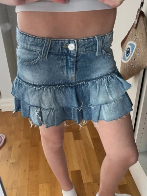 Blå volangkjol i jeans från Gina Tricot - Supersöt kort jeanskjol från Gina Tricot med två volanglager och råa kanter. Kjolen har klassisk femficksdesign, knapp och dragkedja framtill. Perfekt för dig som gillar en avslappnad men trendig stil.