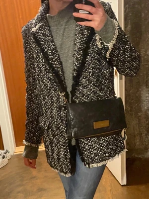 Svartvit och silver bouclé kappa med fransar - Stilren svartvit bouclé kappa med fransiga kanter och markerade slag. Kappan har en rak passform och är tillverkad i ett grovt, texturerat tyg som ger en trendig look. Perfekt för dig som vill ha en chic och modern ytterjacka. Glittrar jättefint