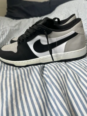 Nike Air Jordan 1 Low svart/vit/grå - Snygga Nike Air Jordan 1 Low sneakers i svart, vitt och grått läder. Klassisk siluett med svart swoosh på sidan och Air Jordan-logga på hälen. Skorna har svarta skosnören och vit mellansula. Perfekta för dig som gillar streetwear och ikoniska sneakers.
