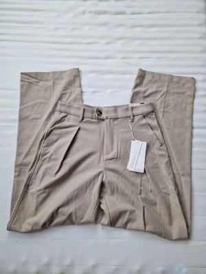 kostymbyxor från bershka - Stilrena beige kostymbyxor från bershka med vida ben och hög midja. Byxorna har pressveck, sidofickor och hällor för skärp. Tillverkade i ett mjukt, vävt material som ger en snygg och avslappnad look. Perfekta för dig som gillar minimalistisk stil.