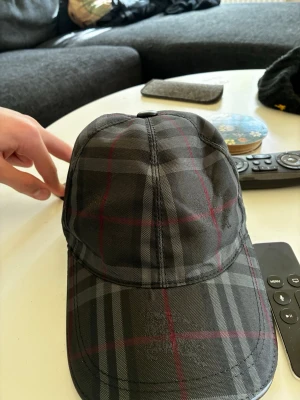 Burberry keps i mörk tartan - Snygg keps från Burberry i mörkgrått tartanmönster med tunna röda och svarta ränder. Klassisk böjd skärm och justerbar passform. Tillverkad i ett slitstarkt tyg som känns lyxigt och trendigt. Perfekt accessoar för dig som gillar exklusiv streetwear.