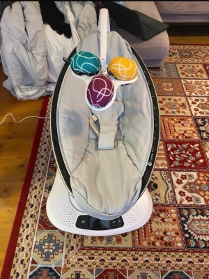Babysitter 4moms MamaRoo grå - Babysitter från 4moms MamaRoo i grått tyg med vit och svart bas. Utrustad med tre färgglada bollar i turkos, gul och lila som hänger ovanför sätet. Modern design och elektronisk styrpanel på foten.