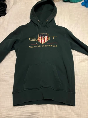 Mörkgrön hoodie från GANT - Snygg mörkgrön hoodie från GANT med broderad logga och texten 'American Sportswear' på bröstet. Klassisk känguruficka framtill, justerbar huva med snören och ribbade muddar. Perfekt för en avslappnad och sportig stil. Bra skick och väldigt stilren hör av er vid mer info 