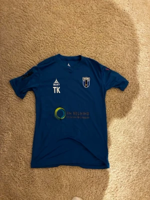 Blå matchtröja från Select - Säljer en blå matchtröja från Select med klubbmärke och flera sponsorloggor både fram och bak. Tröjan har rund hals, korta ärmar och tryckta detaljer i vitt, svart och grönt. Perfekt för träning eller match inom fotboll.