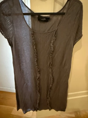 Grå topp med volang från Resteröds - Snygg grå topp från Resteröds med volangdetaljer framtill och rund halsringning. Tunn och lätt i materialet, perfekt för lager på lager. Kortärmad och enkel att matcha till olika stilar.