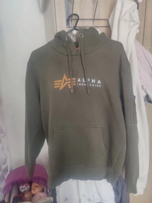 Olivgrön hoodie Alpha Industries - Snygg olivgrön hoodie från Alpha Industries med stor logga framtill, huva med dragsko och magficka. Ärmen har en cool ficka med dragkedja och detaljer. Perfekt för en streetwear-look.