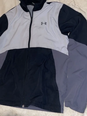 Under Armour vindjacka svart/grå - Snygg vindjacka från Under Armour i svart och ljusgrått med dragkedja framtill och hög krage. Jackan har logga på bröstet och är tillverkad i ett lätt, vindtätt material. Perfekt för dig som vill ha en sportig look.