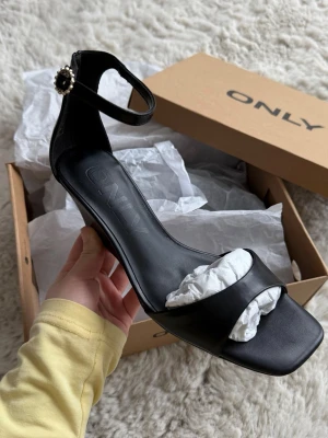 Svarta pumps med dekorativ spänne -  svarta pumps från ONLY med öppen tå och bred rem framtill. Skon har en ankelrem med guldigt spänn, klack på ca 6 cm. Aldrig använd, fortfarande i kartong ⭐️