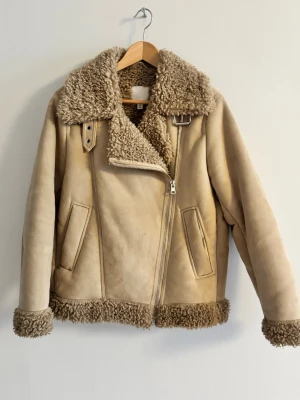 Beige skinnjacka med teddyfoder - Beige skinnjacka med mjukt teddyfoder i krage, ärmslut och nederkant. Jackan har sned dragkedja, två fickor framtill och ett dekorativt spänne vid halsen. Trendig modell som ger en cool look.