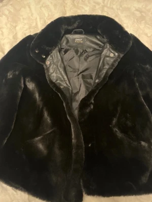 Svart fuskpälsjacka från ONLY - Säljer en svart fuskpälsjacka från ONLY med bred krage och knappar framtill. Jackan har en mjuk, glansig yta och är helfodrad för extra värme. Perfekt för dig som vill ha en stilren och mysig vinterlook.