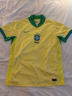 Brasilien Nike fotbollströja gul - Snygg gul Brasilien fotbollströja från Nike med gröna detaljer och blå Nike-logga. Tröjan har korta ärmar, grönt kragparti och diskret mönster i tyget. Perfekt för match eller träning. Officiell CBF-logga på bröstet.