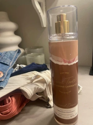 Sweet Tooth Body Mist Caramel Dream - Sweet Tooth Body Mist från Sabrina Carpenter i doften Caramel Dream. Flaskan är cylindrisk och genomskinlig med guldfärgad spraypump och lock. Designen har bruna och vita toner med elegant text. Innehåller 236 ml och ger en söt, karamellaktig doft.