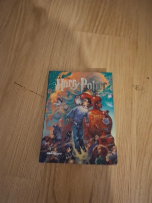 Harry Potter bok med färgstarkt omslag - En Harry Potter-bok med ett färgstarkt och detaljerat omslag där Harry svävar i luften omgiven av magiska element och karaktärer. Boken har mjukt omslag i papper och illustrationerna är livfulla med inslag av blått, orange och rött.